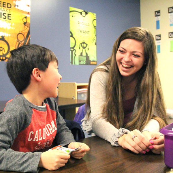 A younger male student with laughing with an older female student at a table in a classroom.